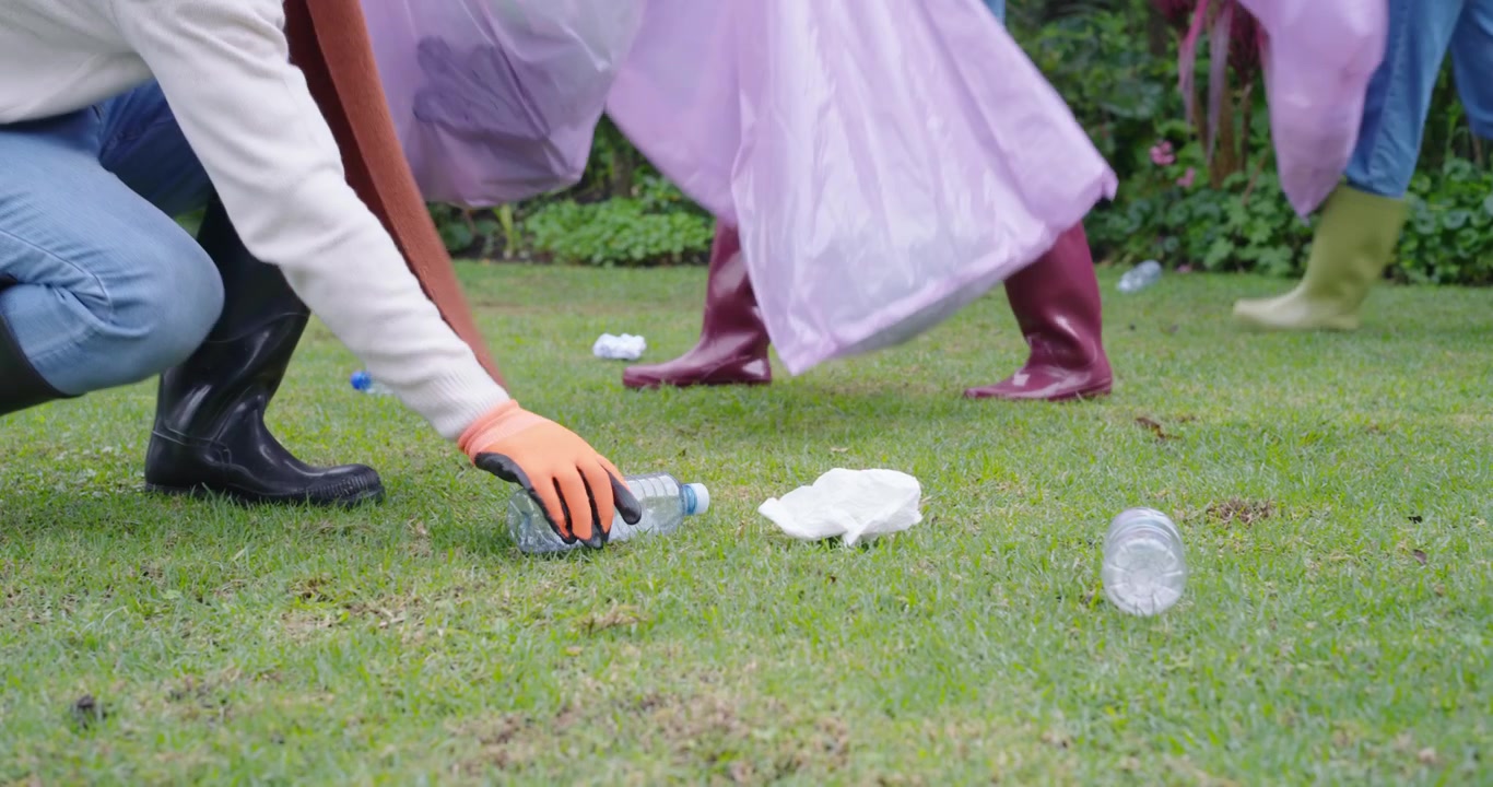 Diverse volunteers entering with pink bags, kneeling, picking bottles in boots and gloves on lawn