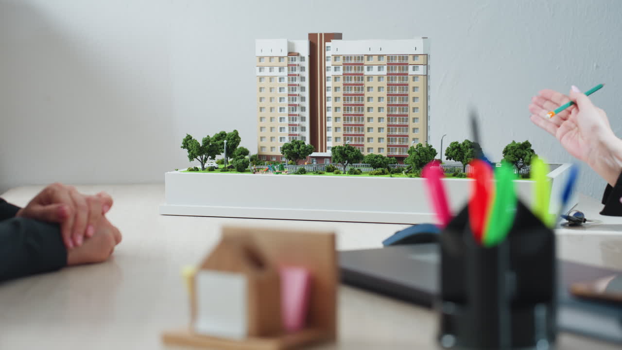 Business expert pointing pencil at modern residential building model while explaining project details to client during office meeting, with green miniature trees and desk items in foreground