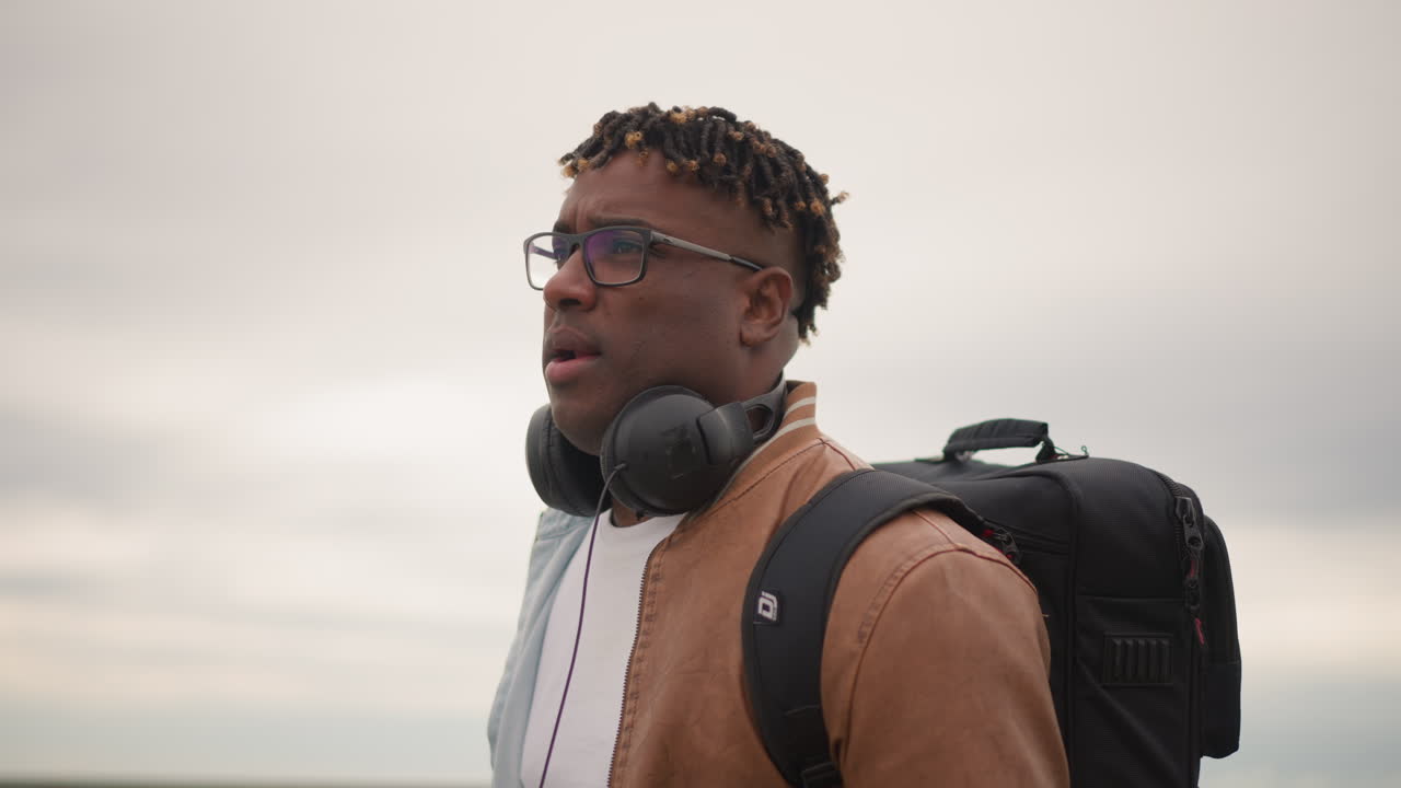 Man with Headphones Checks Phone Amid Cityscape, Male Individual with Glasses and Backpack Prepares Music Session, Enthusiastic Urban Musician in TelevisionStyle Attire Gets Ready for Performance