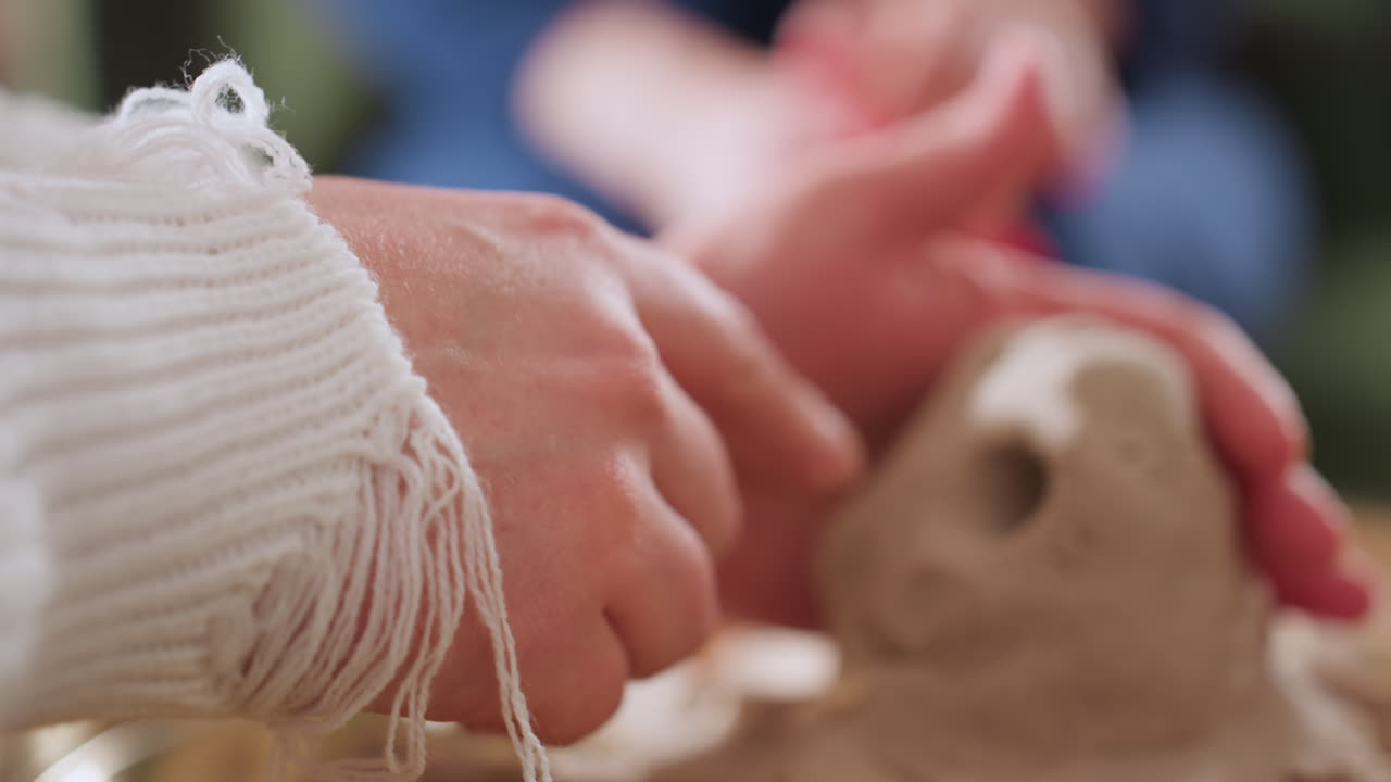 Hands of vulnerable visitor carefully shaping octopus figure in tray during therapy session, soft focus on background figure in blue gown creates emotional atmosphere and sense of shared presence