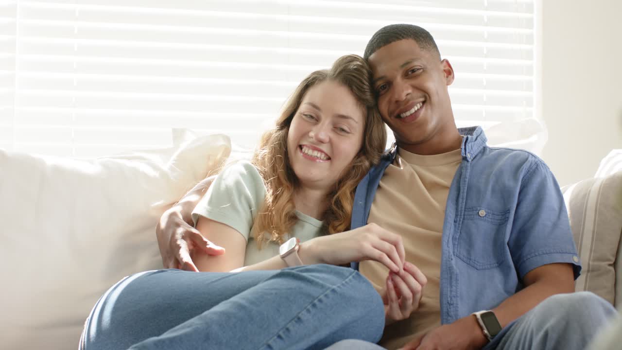 Diverse couple reacting to man's smile by holding hands, leaning on sofa at home sharing affection