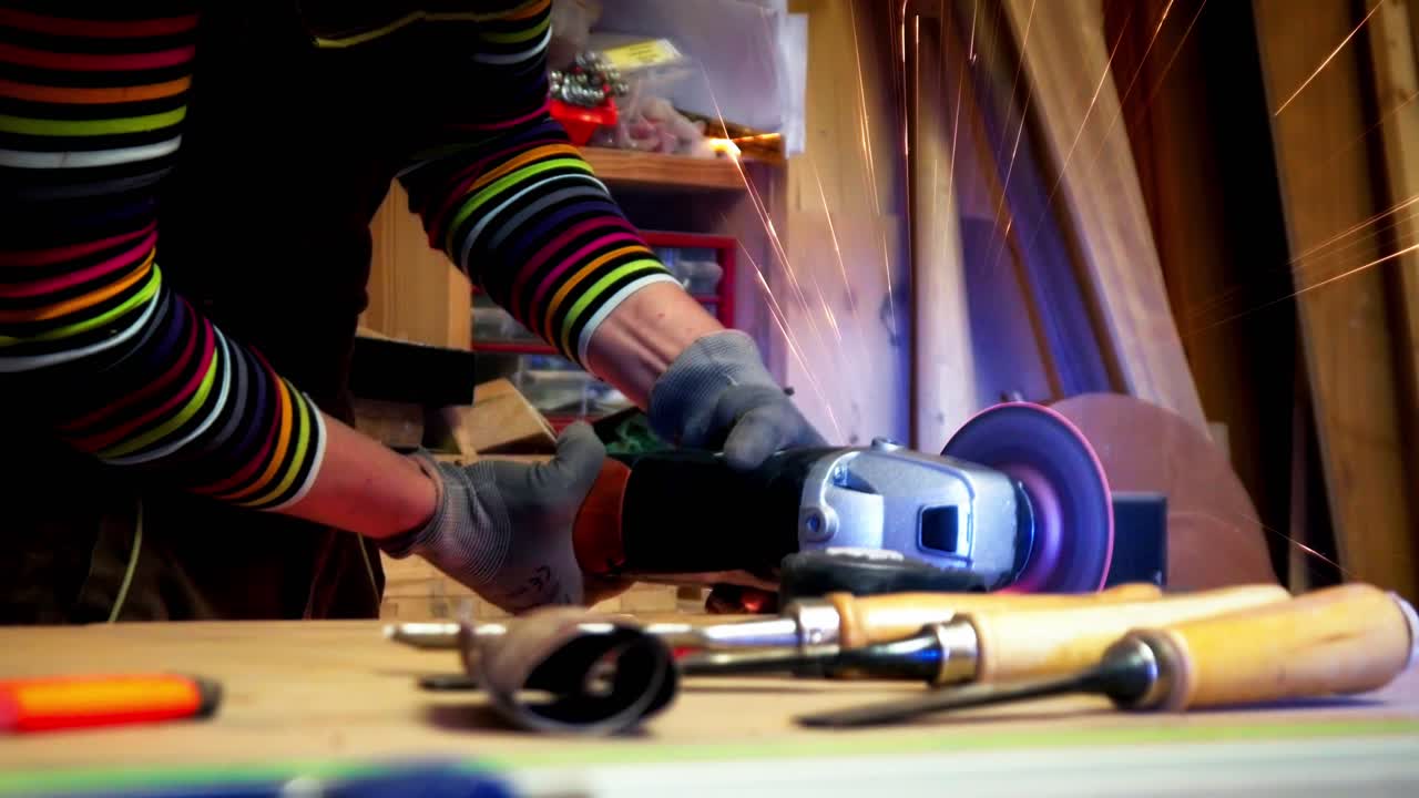 Artisan woman using an angle grinder to cut metal on a workbench, creating a shower of sparks in her home workshop. Close-up shot