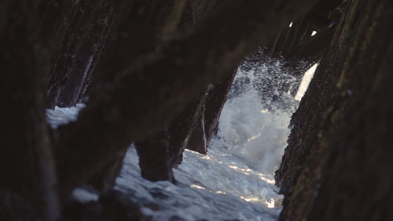 olas rompiendo debajo de un muelle, viejos pilotes de madera con rayos de sol que atraviesan