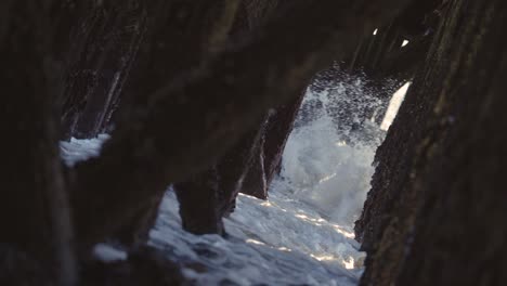 Waves-crashing-under-a-pier,-old-wooden-pillings-with-sunbeams-coming-through