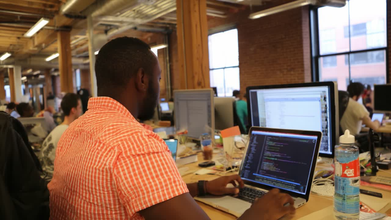 Focused Programmer Engaged in Coding Tasks Inside a Modern Open-Office Space, with Multiple Computers and Colleagues Working Nearby