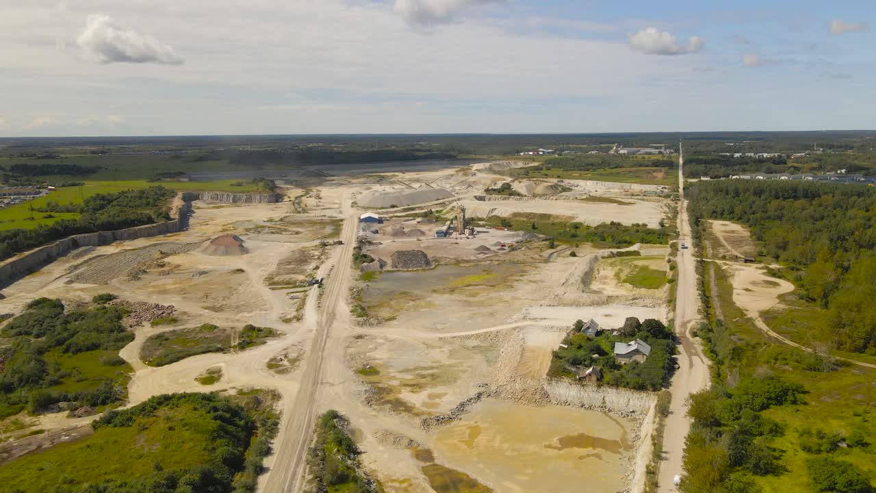 Flying above vast open pit quarry mining site of limestone materials extraction, overburden piles and dust roads. Exposed rock layers, surface mining for construction industry under cloudy summer sky