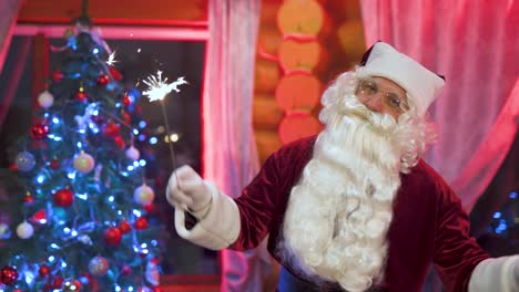 happy santa claus is dancing near the christmas tree with sparklers. house with new year's decorations on the eve of the holiday