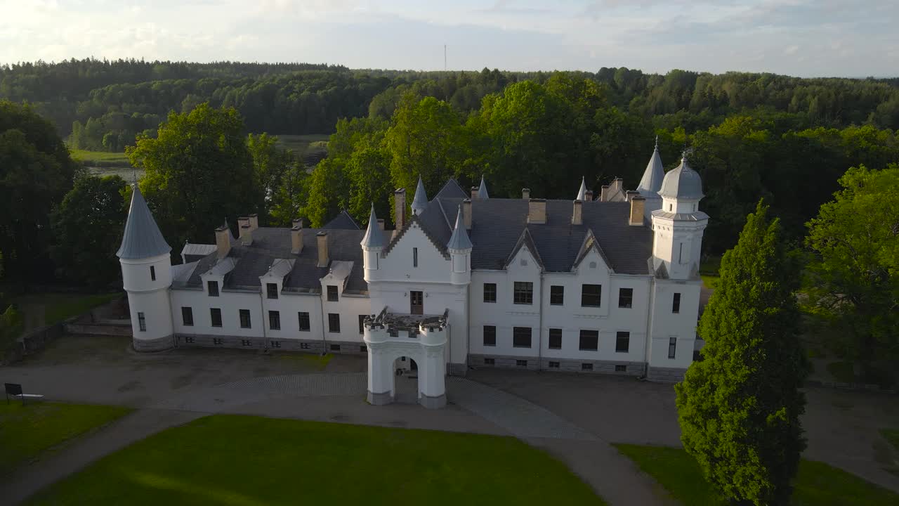 Gorgeous aerial view ascending in front of Alatskivi white colored majestic castle during a sunny summer day with green mowed grass in front of it with a road. Blue sky and white clouds behind it.