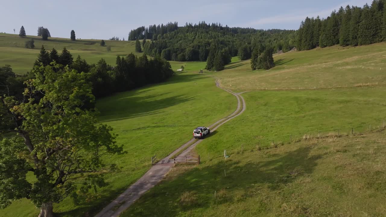 Aerial of a white SUV drives along a winding dirt road through rolling green hills and forested mountains under clear skies, symbolizing freedom, travel, and adventure in nature