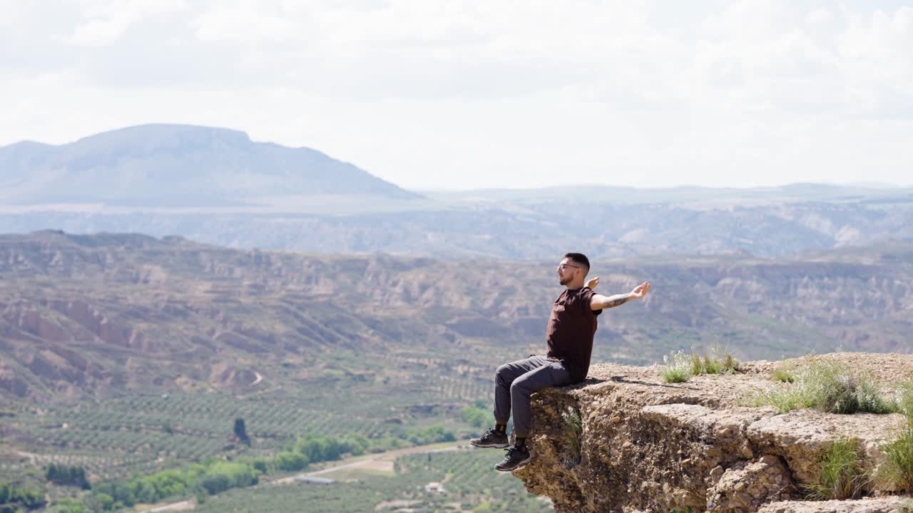 Happy man raising hands sitting on rocky cliff enjoying mountain landscape