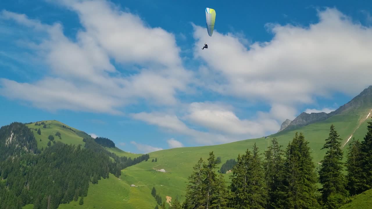 A paraglider glides swiftly against a bright blue sky, with the Fribourg Alps at Schwarzsee, Switzerland behind him and a chairlift passing below
