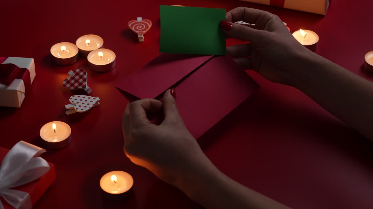 Woman's hands put valentine's postcard with green screen chroma key in envelope. Top view of red table with burning candles, decorative hearts and gift boxes. Romantic twilight. Close up. Slow motion
