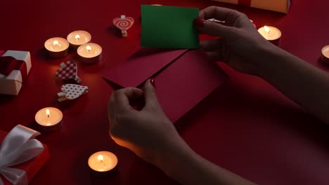 woman's hands put valentine's postcard with green screen chroma key in envelope. top view of red table with burning candles, decorative hearts and gift boxes. romantic twilight. close up. slow motion