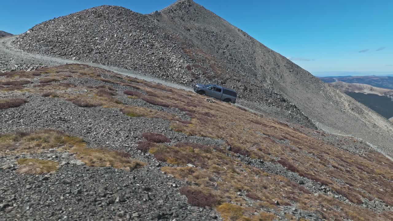 Rearview of offroading truck slowly crawling up steep incline on trail as drone orbits to front view of car with road behind
