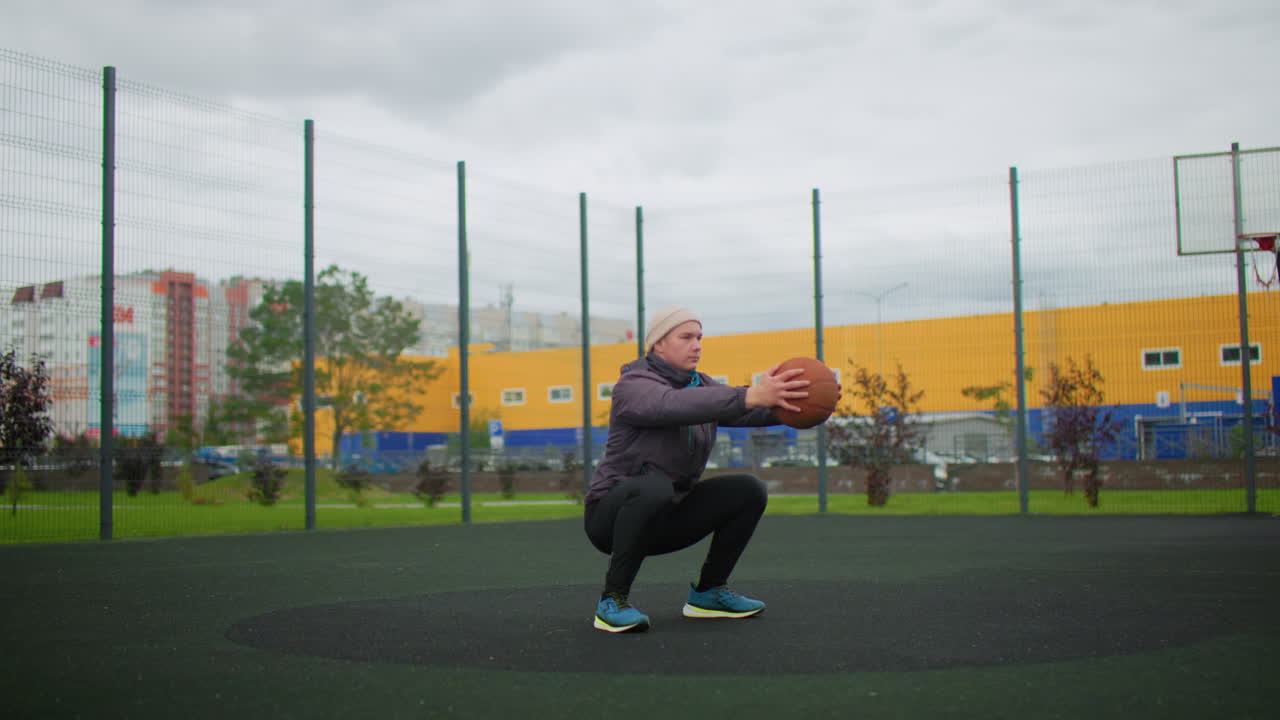 uomo bianco in un campo da basket all'aperto che tiene una palla durante una fredda sessione di allenamento urbano, berretto e giacca, esercizio di squat, postura concentrata, edificio industriale giallo e recinzione metallica sullo sfondo