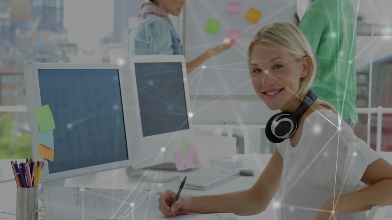 Businesswoman at desk checking monitors and writing as team adding sticky notes overlay over desk