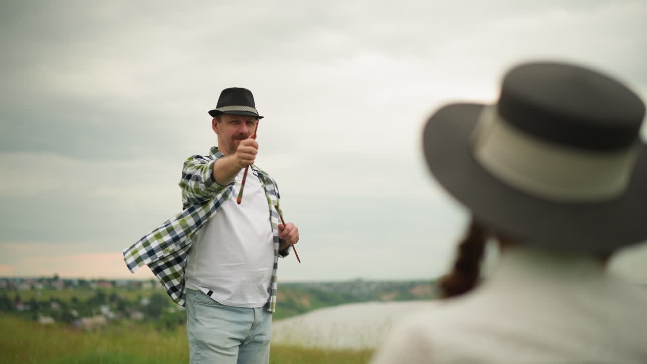 체크 된 셔츠를 입고 을 들고 웃는 장인, 배경에 모자를 입은 흐릿한 여자를 가리키고, 평온한 풍경과 함께 야외에서 경쾌하고 창의적인 순간