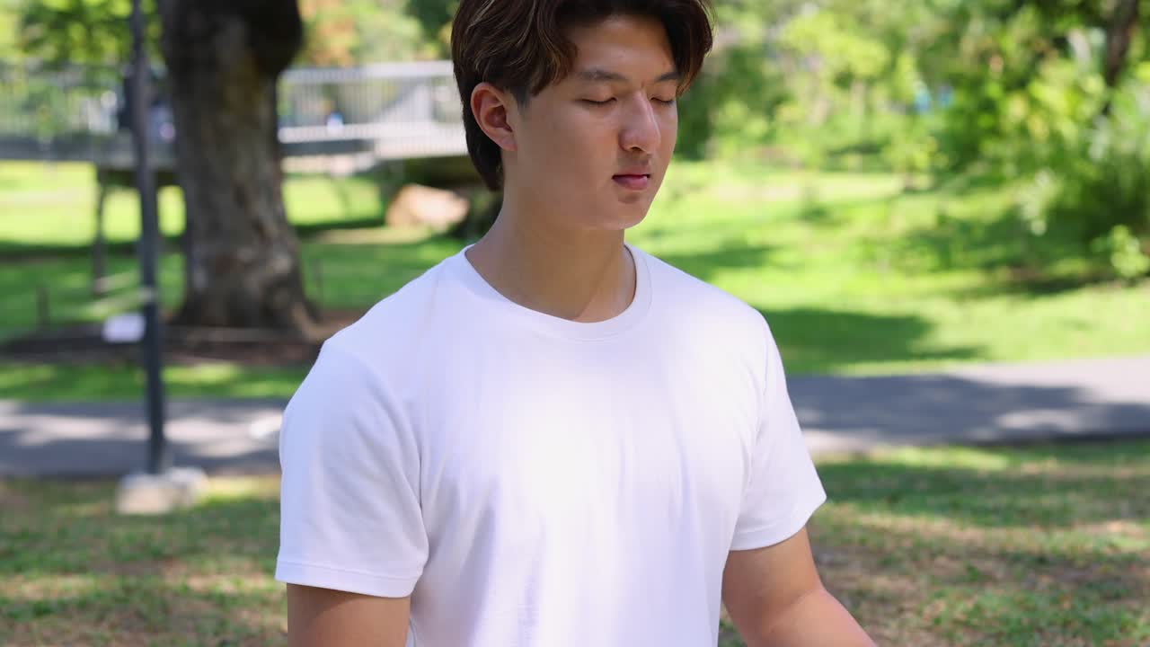 Giovane uomo asiatico pratica respirazione profonda e meditazione mentre è seduto in un rigoglioso parco verde