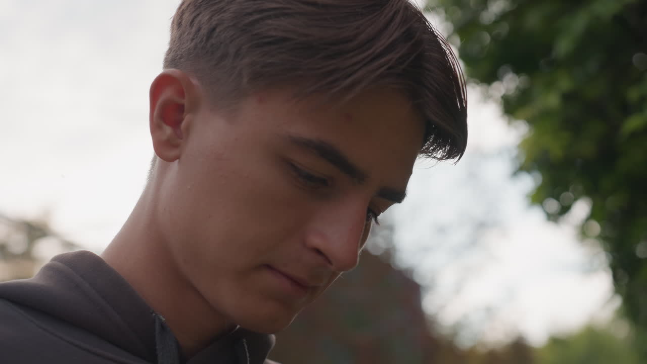Portrait contemplatif en extérieur d’un jeune homme montrant une séquence où il baisse les yeux puis relève le profil, adolescent en sweat à capuche devant un jardin verdoyant, lumière douce et nuageuse, faible profondeur de champ, tons cinématiques, naturel