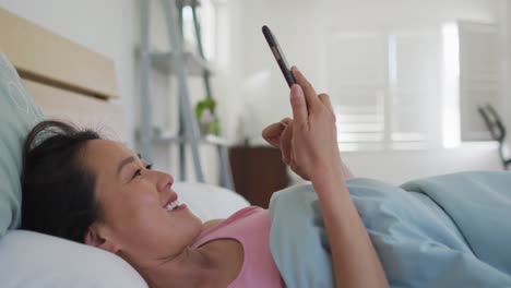 Happy-asian-woman-lying-in-bed,-using-smartphone-in-the-morning