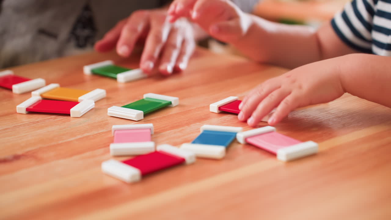 Close up of kid scattering colorful blocks on wooden table while adult hand gathers pieces nearby, striped shirt visible, playful childhood interaction combined with supportive guidance