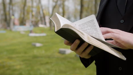 close up view of man hands holding a bible and reading in front of a tombstone in a graveyard 1