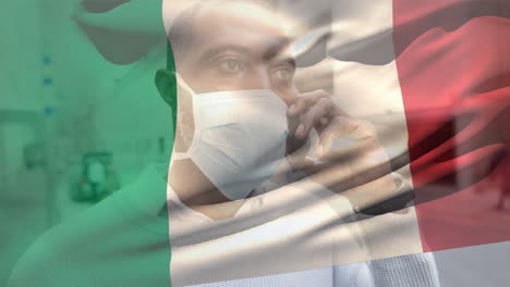 bandera italiana ondeando contra el hombre que lleva la máscara de la cara hablando en el teléfono inteligente