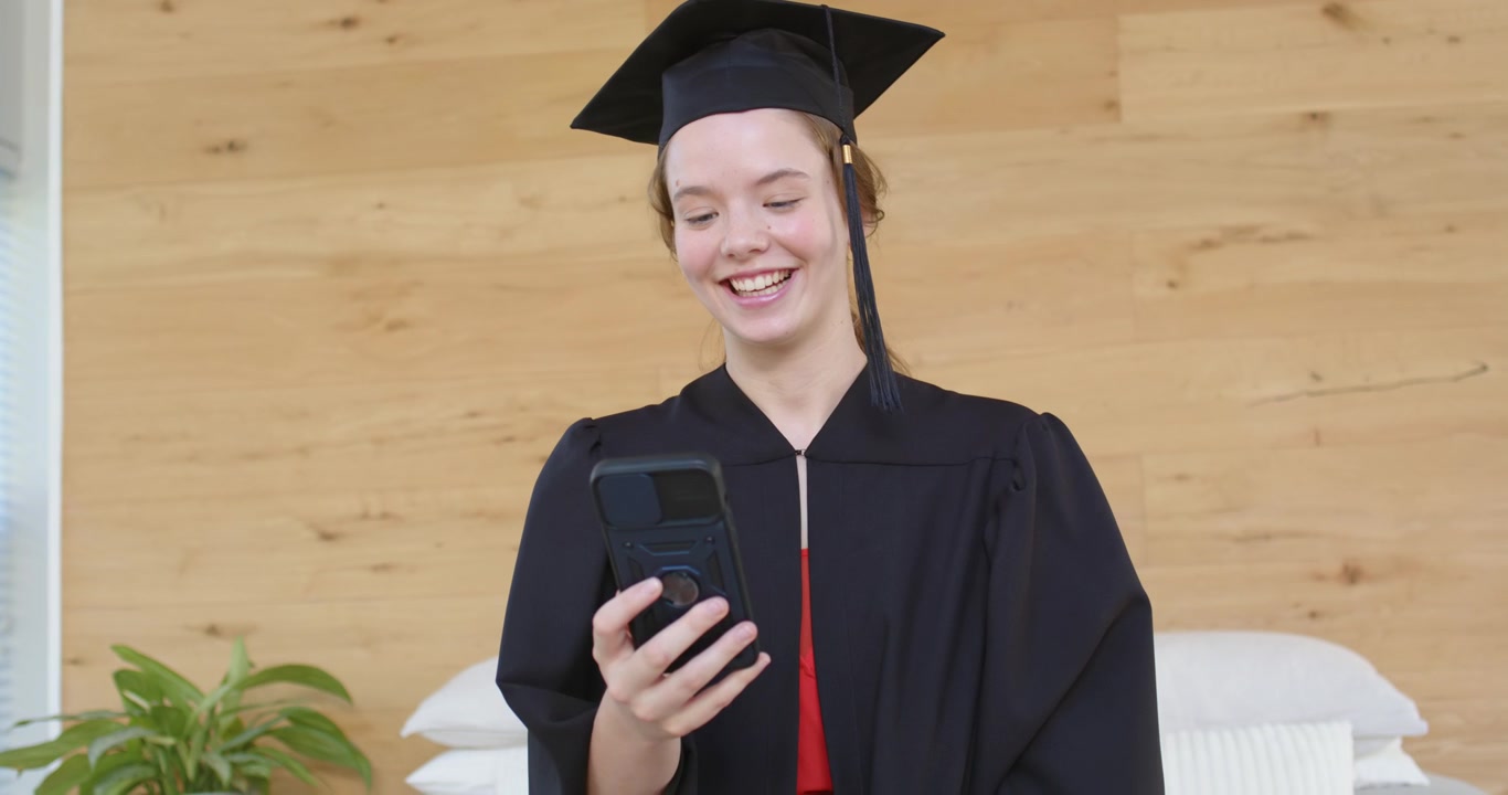 Donna adulta in toga che riceve una chiamata per la laurea, tiene il telefono a casa, saluta e sistema il fiocco del tocco.