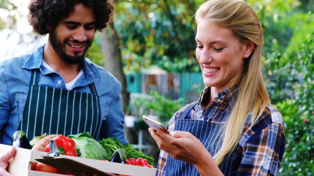 hombre llevando una caja de verduras mientras la mujer habla por teléfono móvil