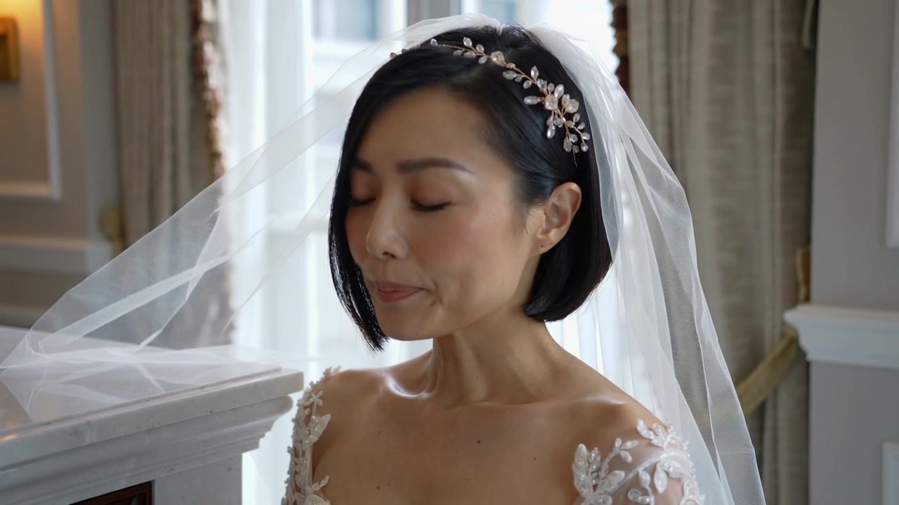 宝石のヘッドピースとレースのガウンを身につけた花嫁が窓辺に立ち、そよ風がベールを揺らす中、誓いのために身構えている。