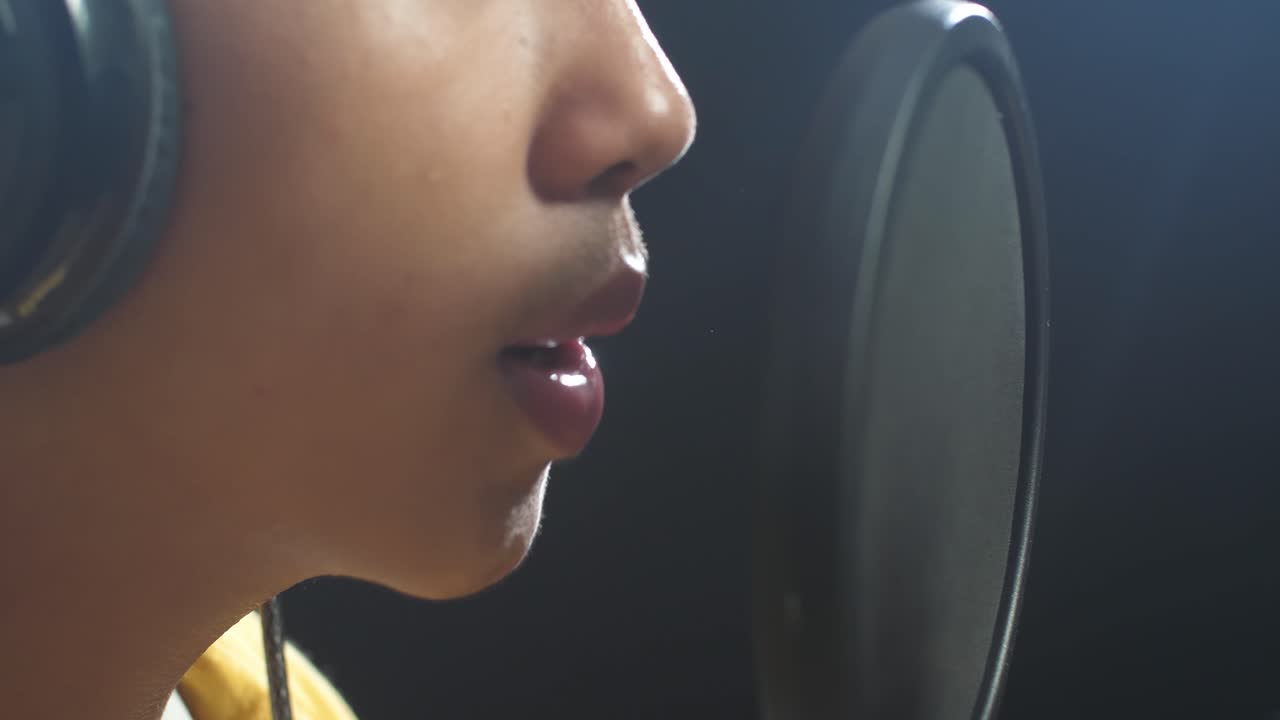 젊은 아시아 소년의 입이 검은 바탕에 있는 콘덴서 마이크로 노래하는 근접한 측면 시각