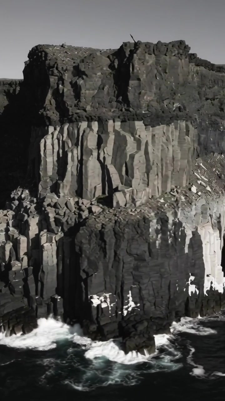 Vertical video: Panning camera revealing tall basalt column cliff at rocky shore, waves crashing