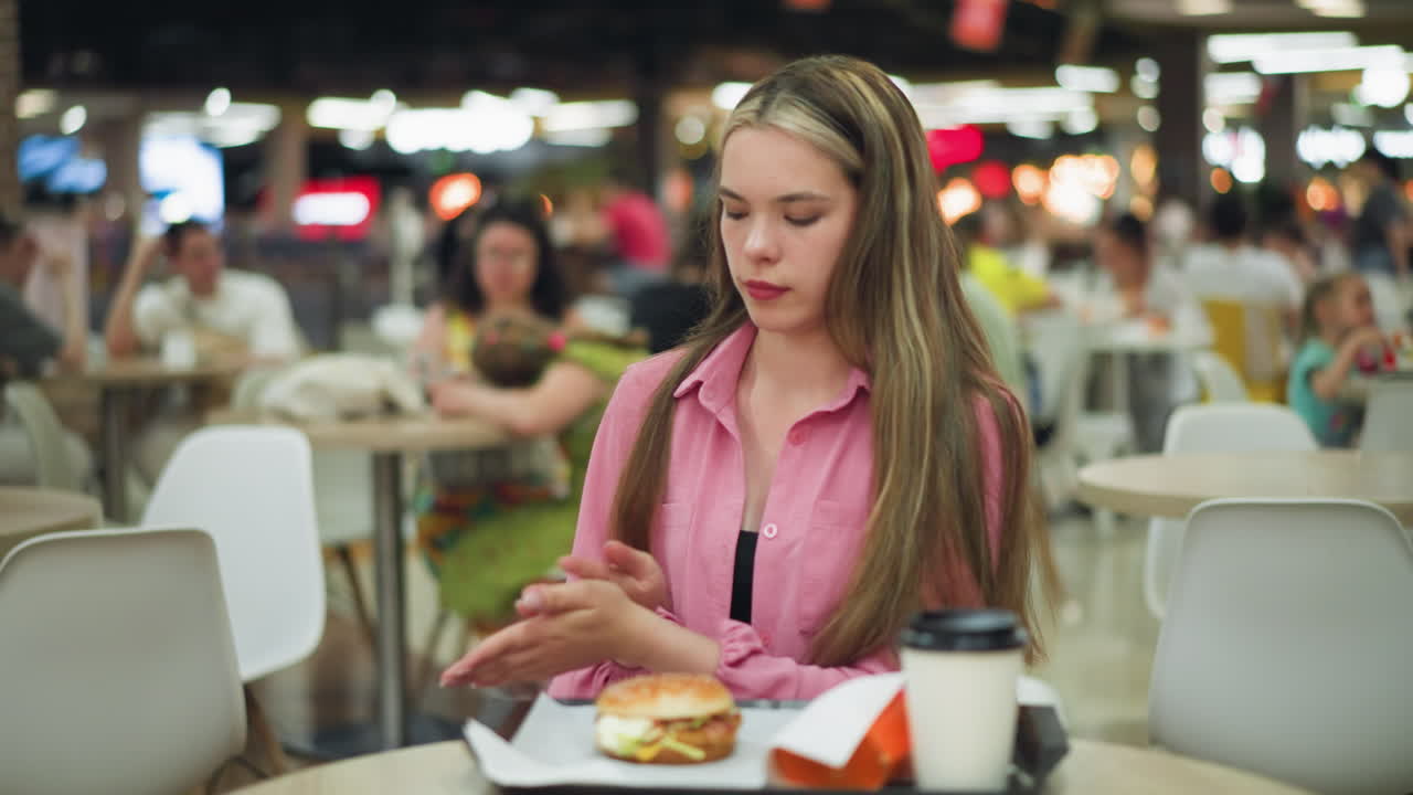 hermosa mujer en vestido rosado limpia las manos, sentada en una mesa de restaurante con hamburguesa, papas fritas y café delante de ella, después de la limpieza, se acerca a la hamburguese, con una vista borrosa de la gente