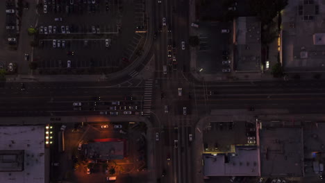 AERIAL:-Birds-eye-view-of-Culver-City,-Los-Angeles,-California-traffic,-intersection-at-dusk-with-car-traffic-passing-and-parking-lot