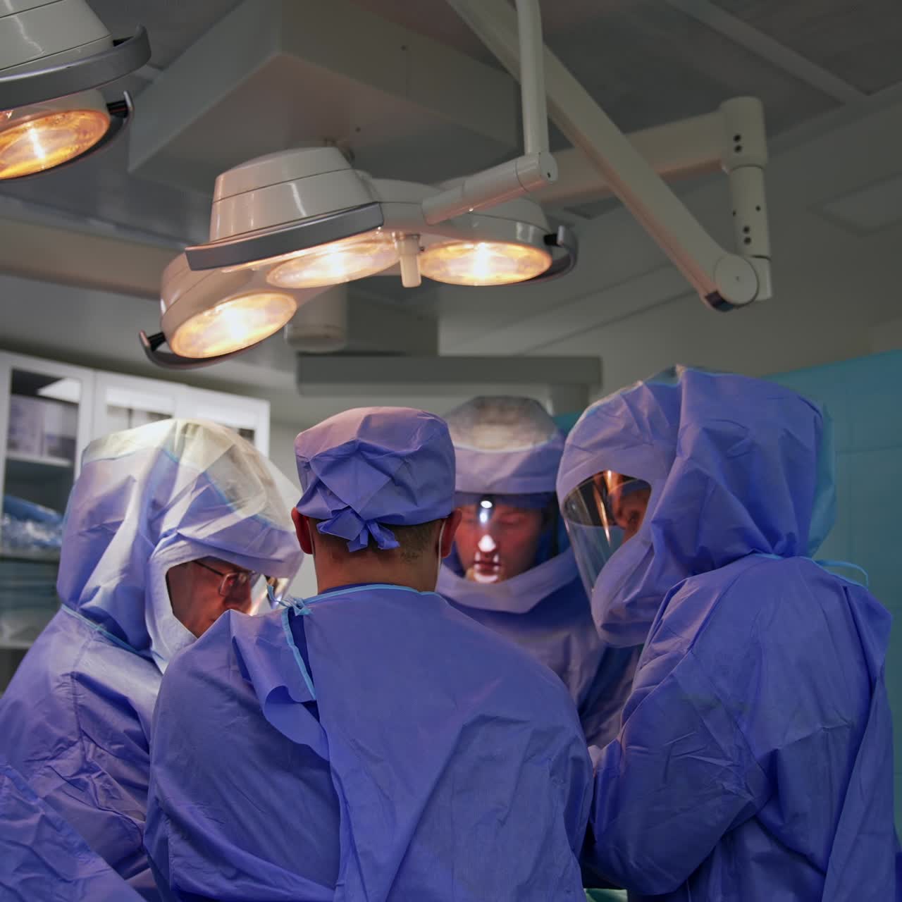 Surgeons wearing special protective suits stand around the operational table. Female nurse giving the instruments to the doctors