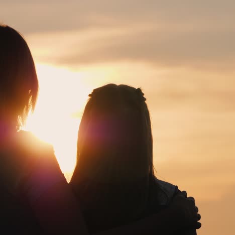 a woman gently hugs her daughter watching the sunset together