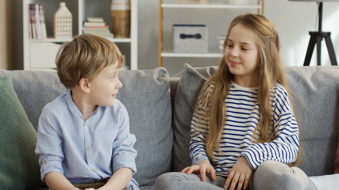 Portrait Shot Of The Cute Small Sister And Brother Sitting On The Couch In The Living Room, Hugging Each Other And Posing In Front Of The Camera