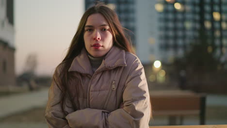 una chica con una chaqueta de melocotón con las manos cruzadas sobre sus brazos se encuentra en una suave luz de la tarde, con un fondo urbano borroso