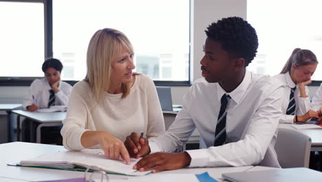 high school tutor giving uniformed male student one to one tuition at desk in classroom