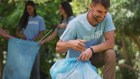 hombre caucásico sonriente y un grupo diverso de amigos poniendo basura en bolsas de basura azules en el parque