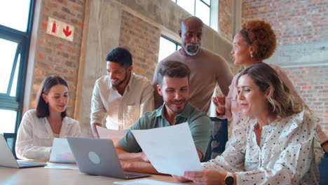 multi-cultural business team meeting around laptop discussing documents in busy office