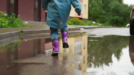 niño feliz corre a través del charco en la calle de cerca niño sano en chaqueta y botas impermeables juega