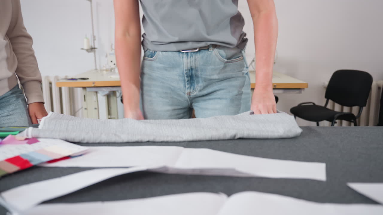 Lower view of dress stylist carefully folding apparel over colorful fabric samples in workspace, preparing textiles for fashion project, surrounded by sewing patterns, and tailoring tools