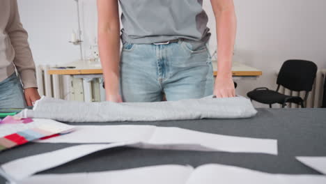 lower view of dress stylist carefully folding apparel over colorful fabric samples in workspace, preparing textiles for fashion project, surrounded by sewing patterns, and tailoring tools