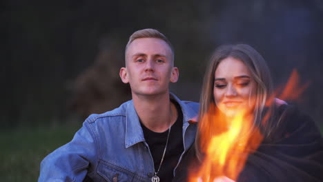 happy young couple sitting and hugging near the campfire and talking. dating pair holding barbecued marshmallows.