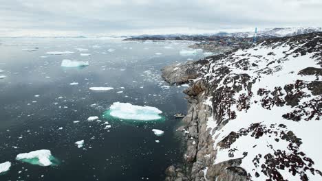 rocky coast with flowing icebergs and town of ilulissat in distance, aerial view