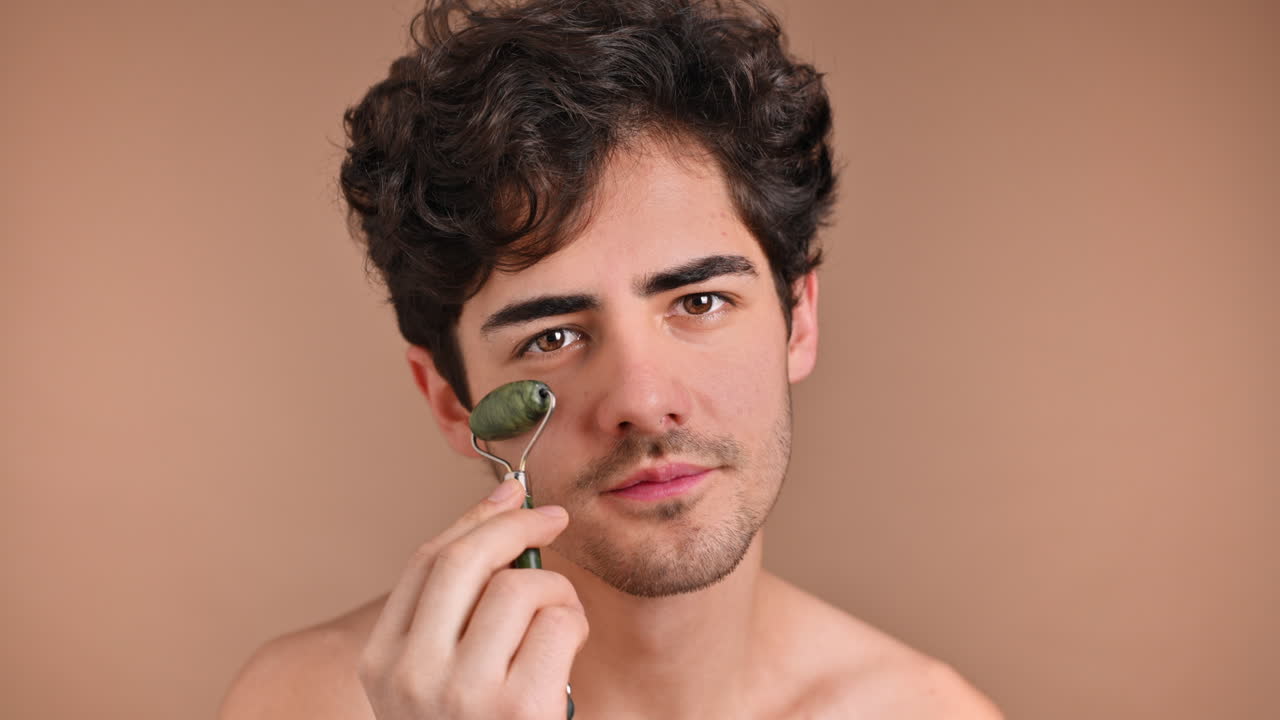 A young caucasian man with stubble beard is doing a facial massage using a roller, looking into the camera