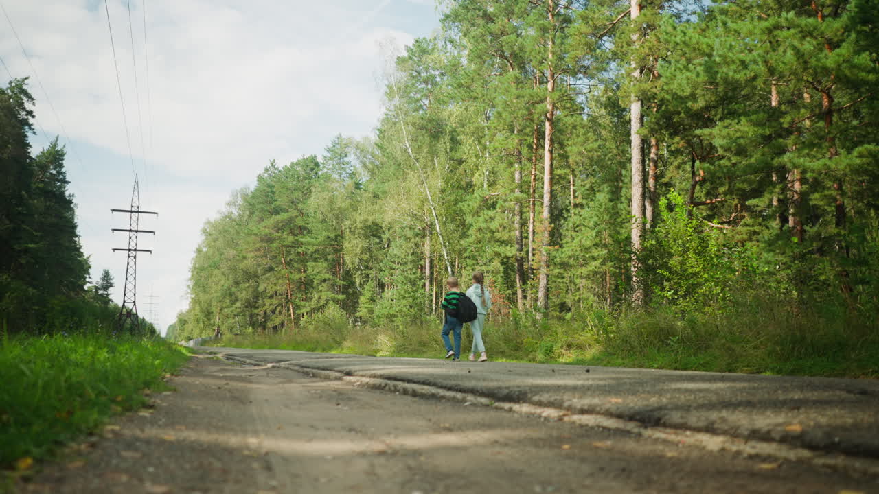 Two children walk beside tarred road surrounded by tall green trees under clear sky, power cables and utility poles stretching across background while low sunlight casts shadows on broken pavement