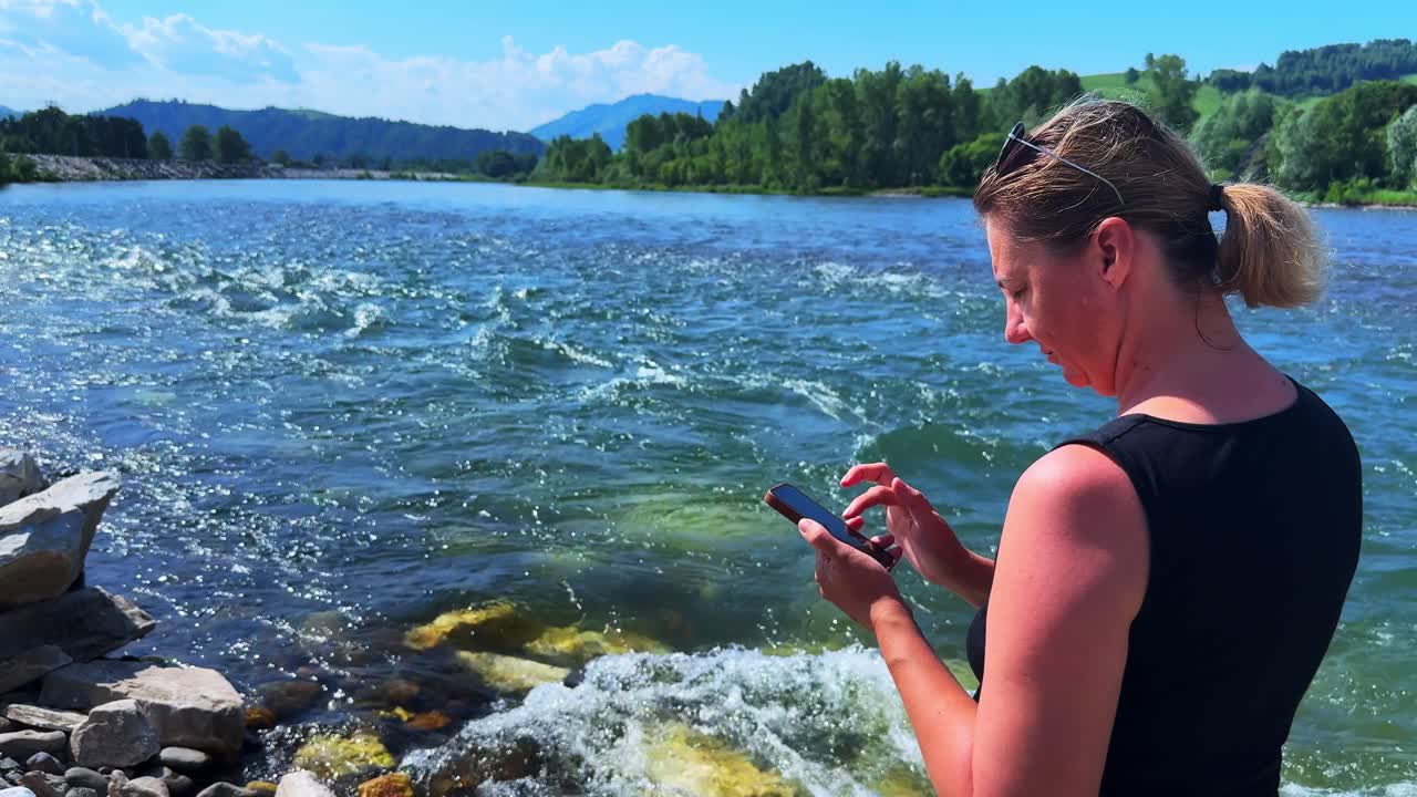 A Woman Engaging with Her Mobile Device by the Serene Riverscape, Enjoying Nature's Beauty While Staying Connected in a Peaceful Outdoor Setting