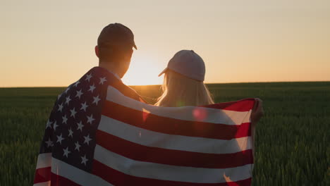 dos agricultores con la bandera de los estados unidos en los hombros miran la puesta de sol sobre un campo de trigo.